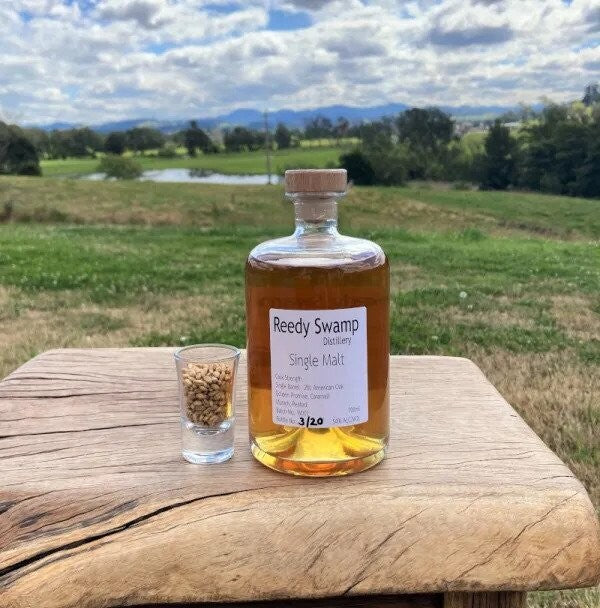 Bottle of Reedy Swamp Single Malt whiskey with a shot glass on a wooden surface with Scenic Bega Valley background