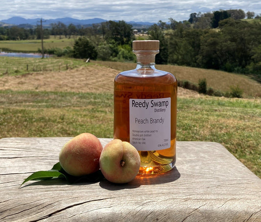 Bottle of Reedy Swamp Peach Brandy with two peaches on a wooden surface outdoors.